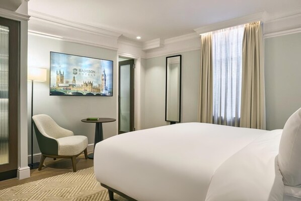 Ein Hotelzimmer mit einem großen Bett, einem Sessel, einem kleinen Tisch, einem Stadtbild als Gemälde und einem Spiegel.