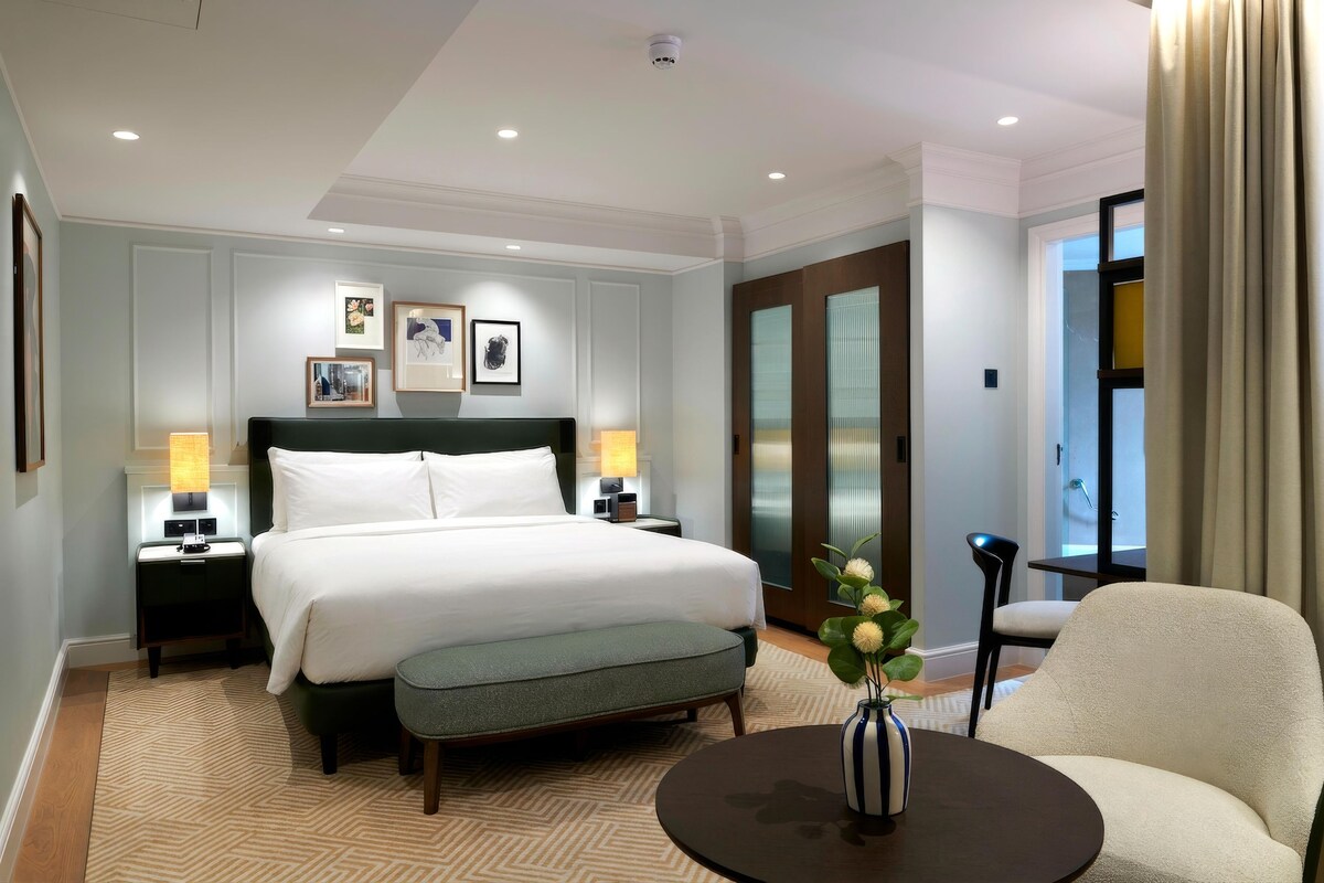 A hotel room with a large bed, bedside tables, a desk, a chair, a vase with flowers, and a view of the outside.