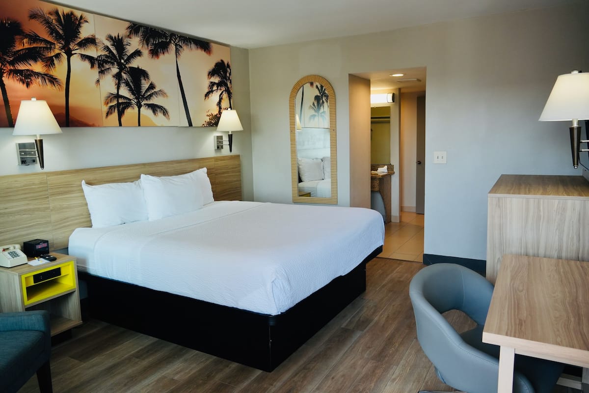 A hotel room with a large bed, bedside lamps, a desk, a chair, a mirror, and a wall with a palm tree painting.