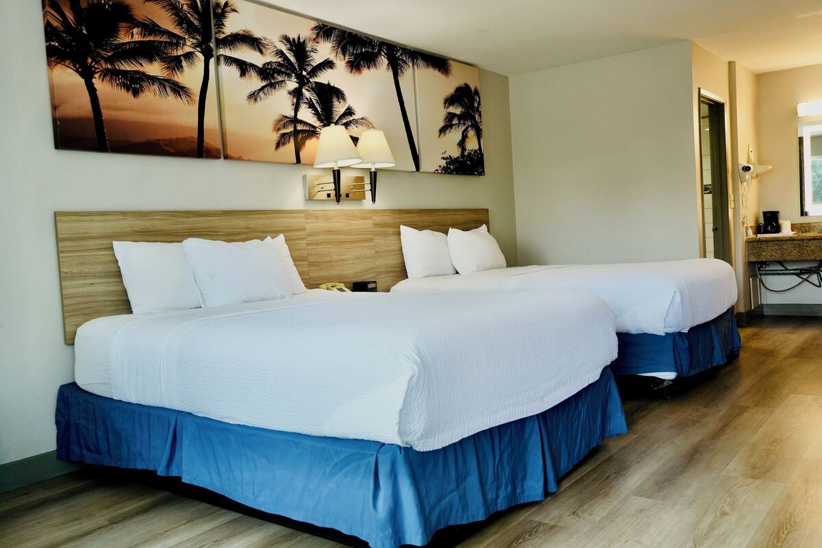 A hotel room with two beds, a wooden headboard, a wall-mounted lamp, and a painting of palm trees.