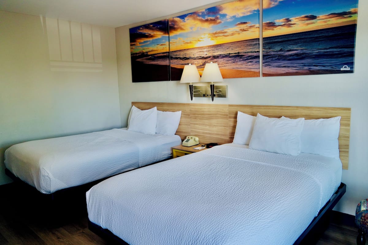 A hotel room with two beds, a wooden headboard, a nightstand with a lamp, and a sunset painting on the wall.