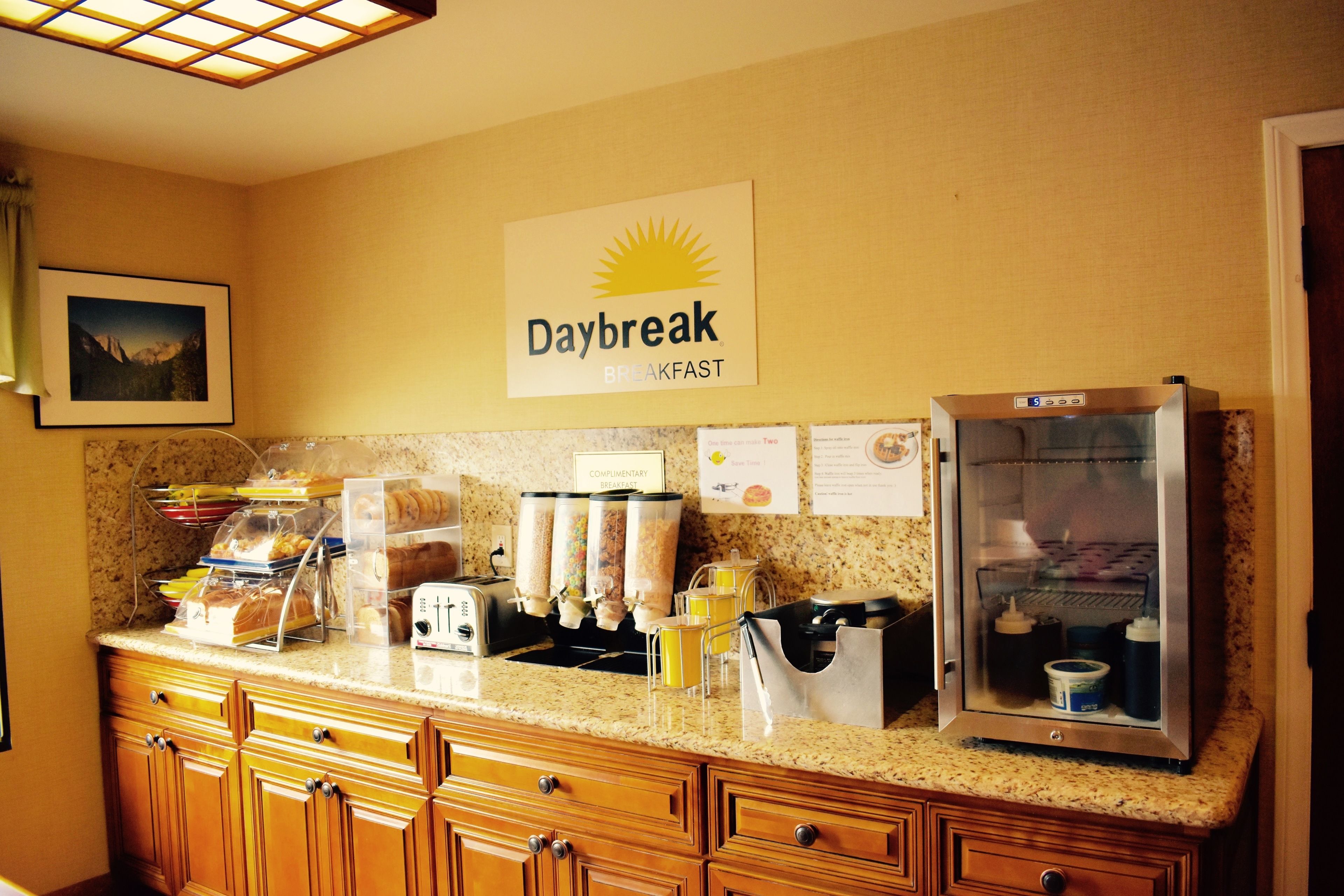 A breakfast area with a sign reading "Daybreak BREAKFAST," a coffee machine, a refrigerator, and various food items on the counter.