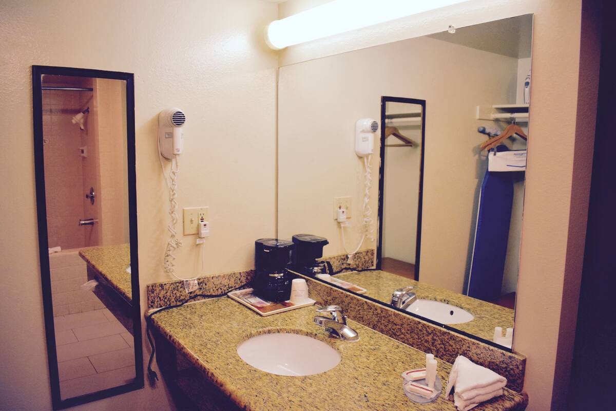 A bathroom with a granite countertop, two sinks, a large mirror, a hair dryer, and a coffee maker.