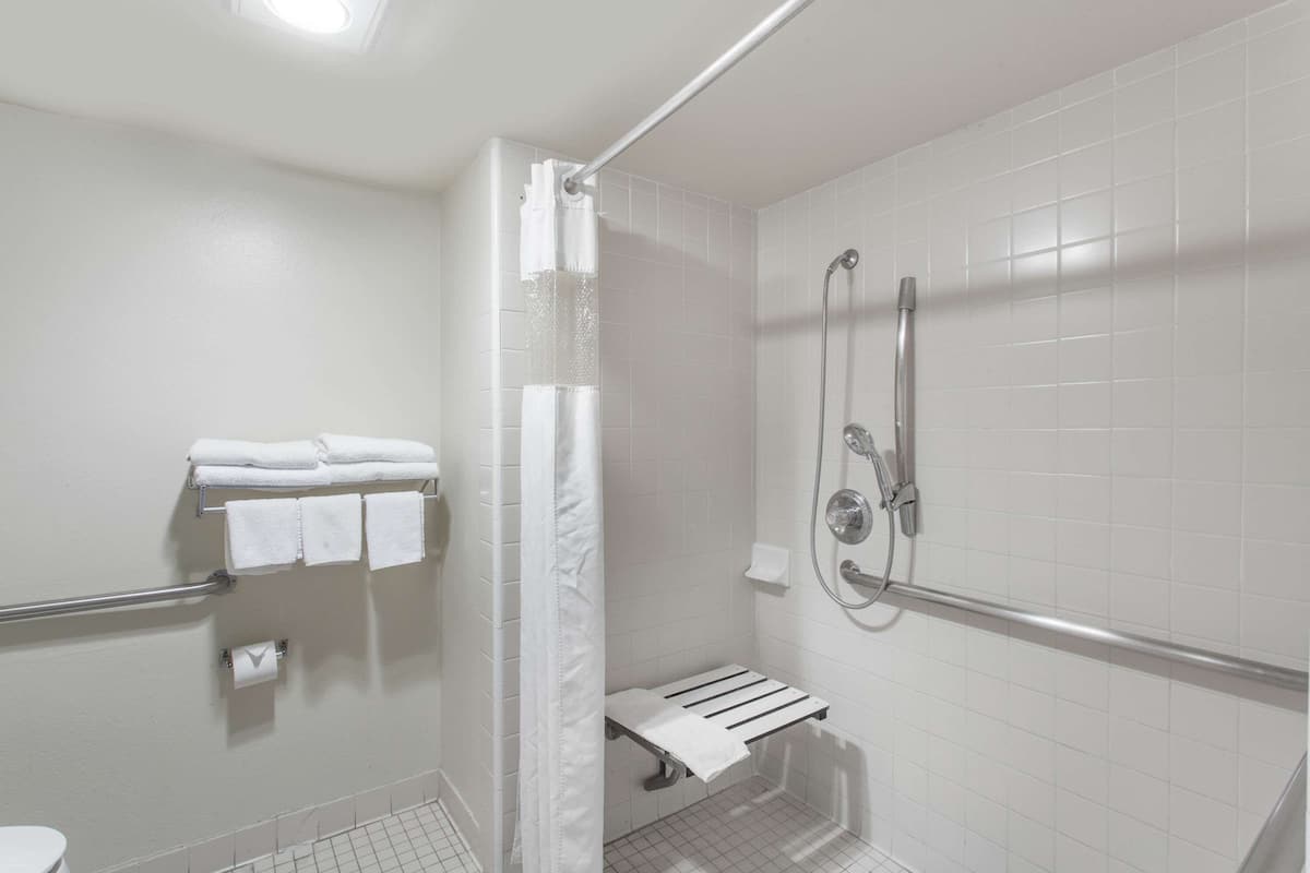 A modern bathroom with white tiles, a shower with a handheld showerhead, a towel rack with folded towels, and a toilet.