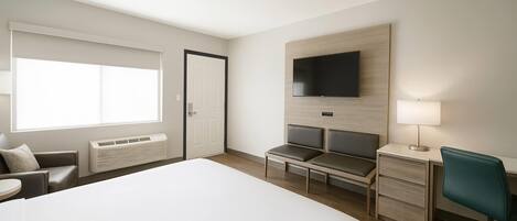 A hotel room with a large bed, a desk with a chair, a television mounted on the wall, a window, and a door.