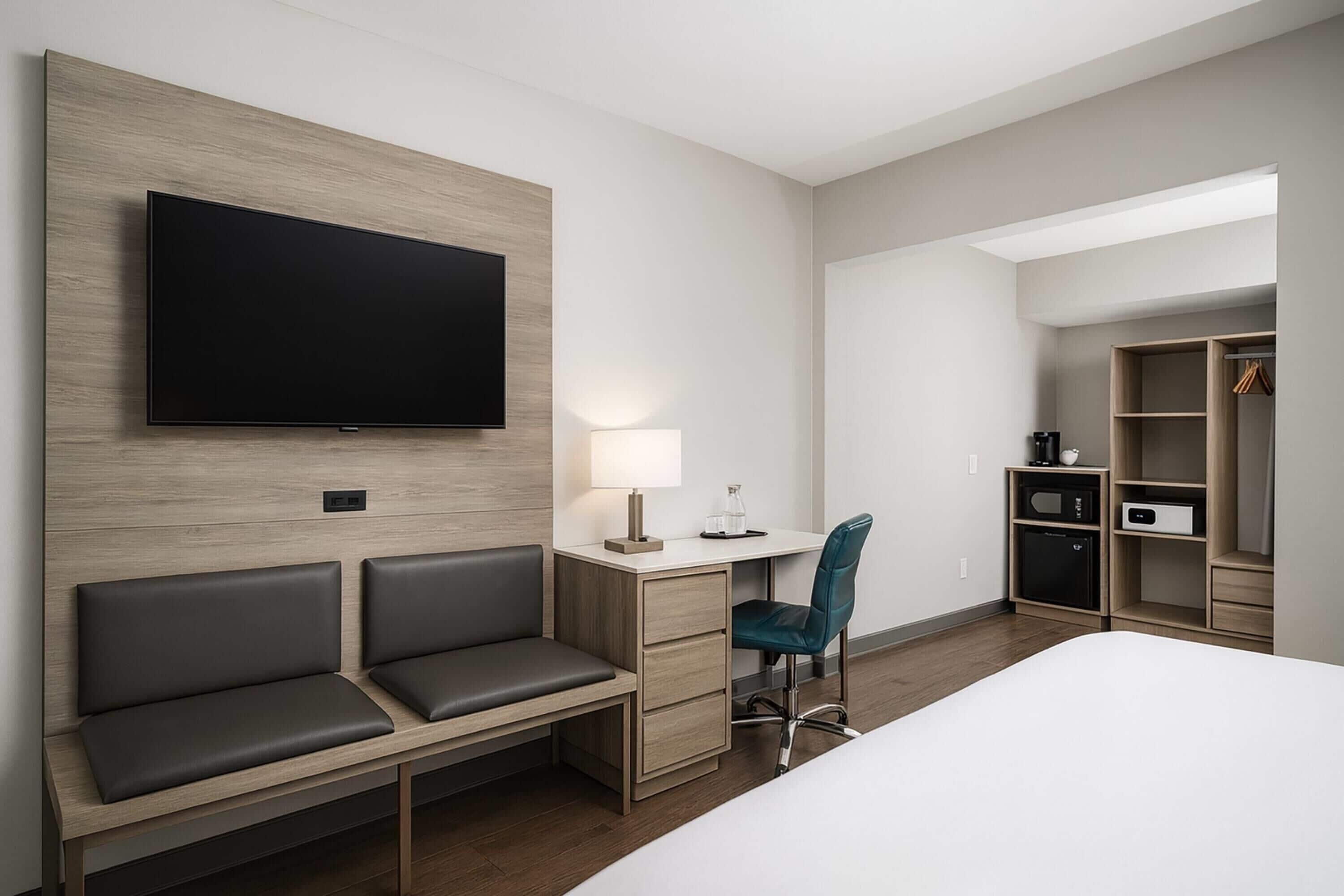 Quarto de hotel com uma cama, uma mesa com cadeira, uma televisão na parede, um abajur e um guarda-roupa.