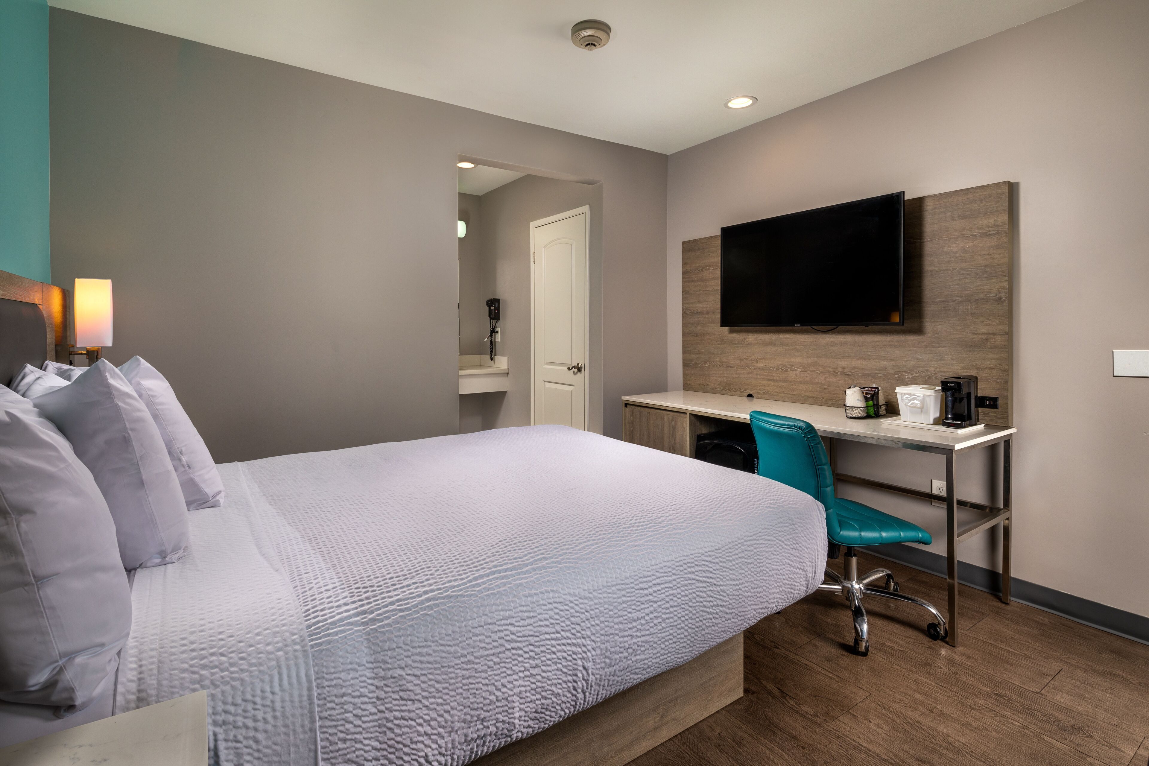 A hotel room with a bed, a desk, a chair, a TV, and a bathroom visible through an open door.