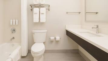 A modern bathroom with a toilet, sink, and bathtub. There are two towels on a shelf above the toilet and a toilet paper roll on the wall.