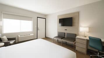 A hotel room with a large bed, a desk with a chair, a TV mounted on the wall, and a window with blinds.
