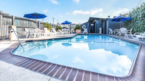 A rectangular swimming pool with a brick edge, surrounded by white lounge chairs and blue umbrellas. - Travelodge by Wyndham LAX (El Segundo)