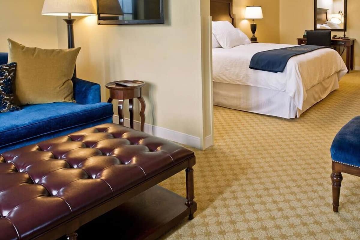 A hotel room with a sofa, a tufted bench, a bed with white bedding, a small round table, and a lamp.