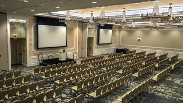 Un salón de conferencias con filas de sillas, un escenario y una pantalla grande.