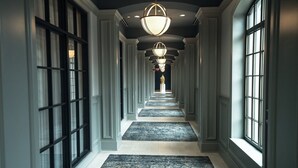 Un pasillo largo y estrecho con alfombra estampada, ventanas amplias a ambos lados y un candelabro en el centro.