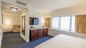 A hotel room with a large bed, a TV mounted on the wall, a wooden dresser, a sofa, and a window with curtains.