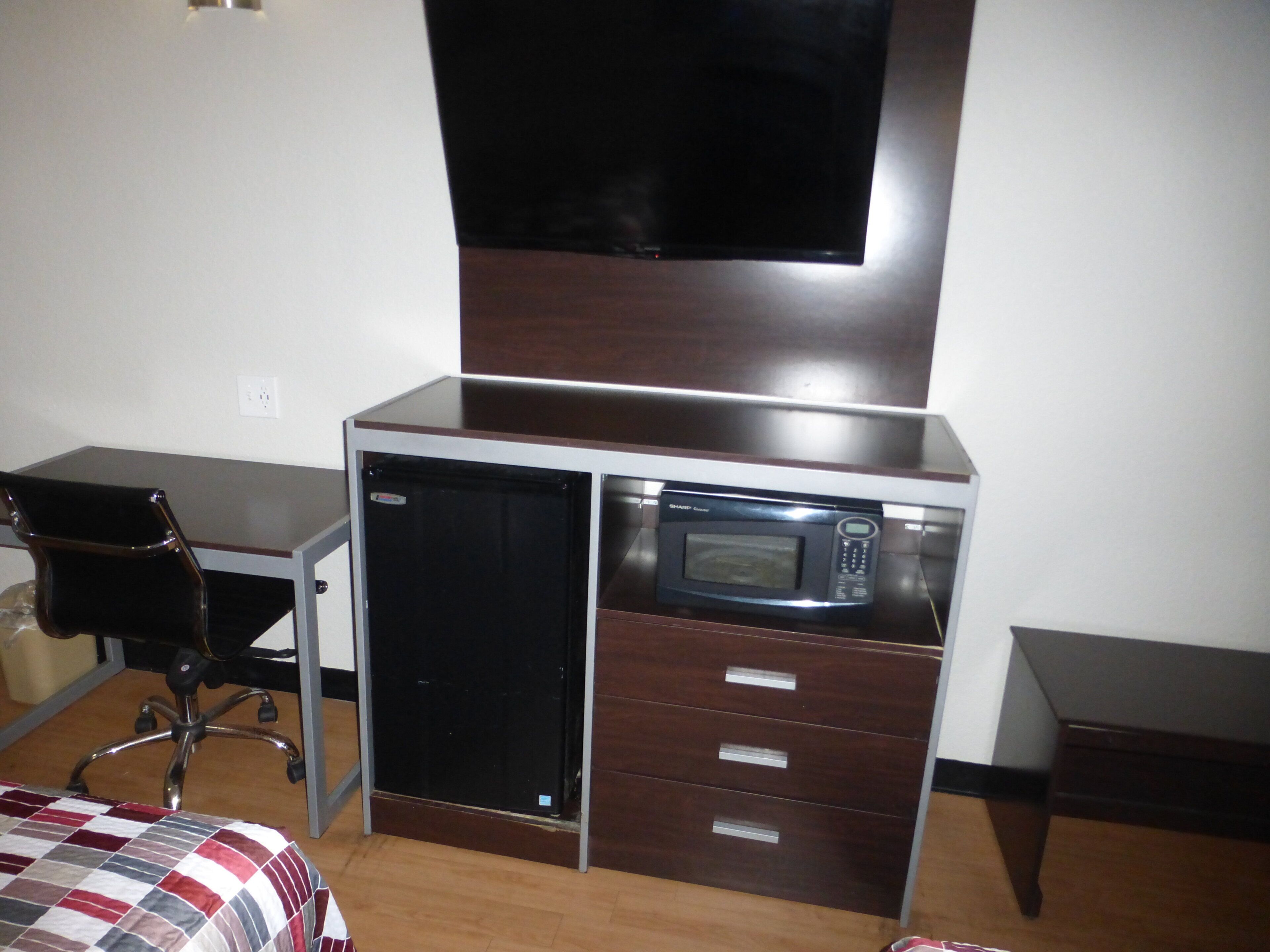 A compact room with a desk, a chair, a bed with a checkered blanket, a microwave, and a television mounted on the wall.