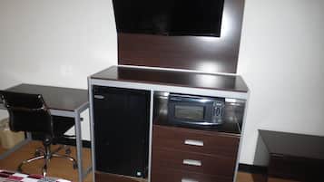 A compact room with a desk, a chair, a bed with a checkered blanket, a microwave, and a television mounted on the wall.