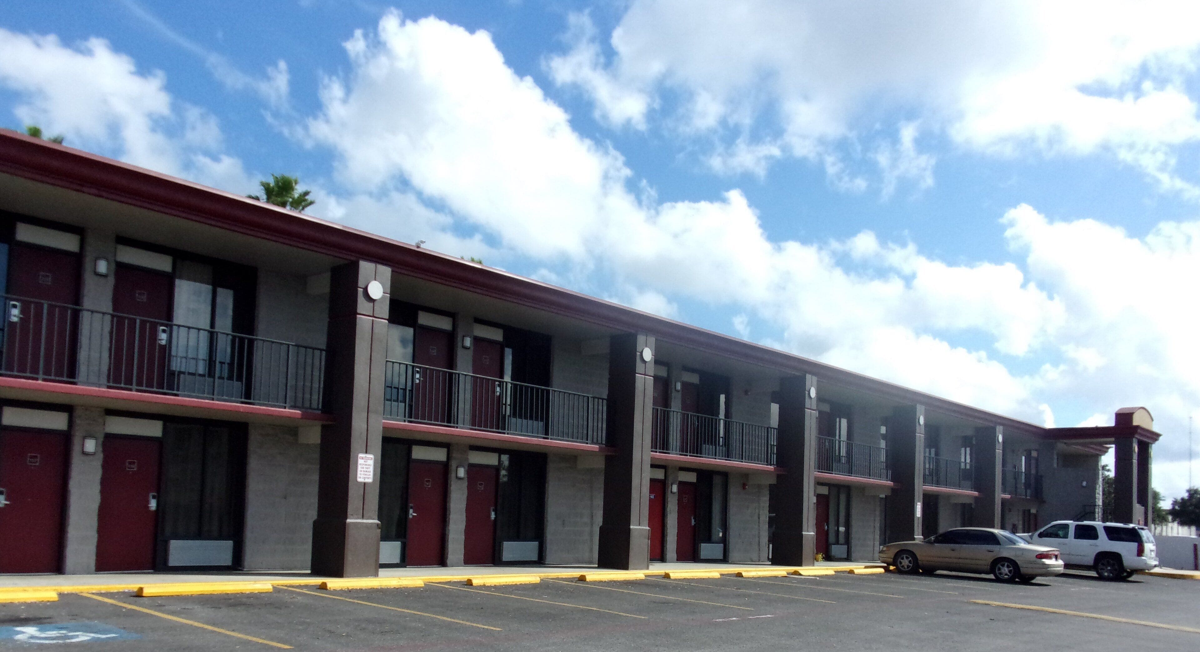 A multi-story motel with a red and beige exterior, multiple balconies, and a parking lot with two cars.