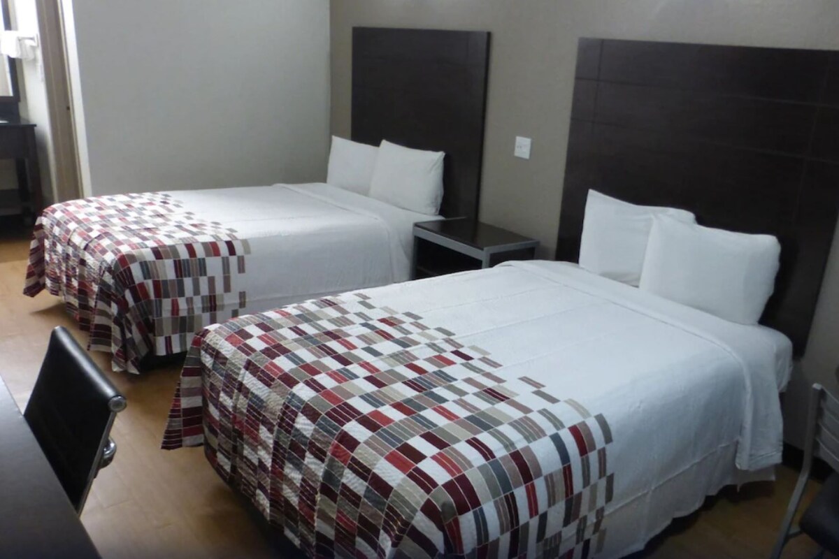A hotel room with two beds, each with a different patterned bedspread. There is a small table and a chair in the room.