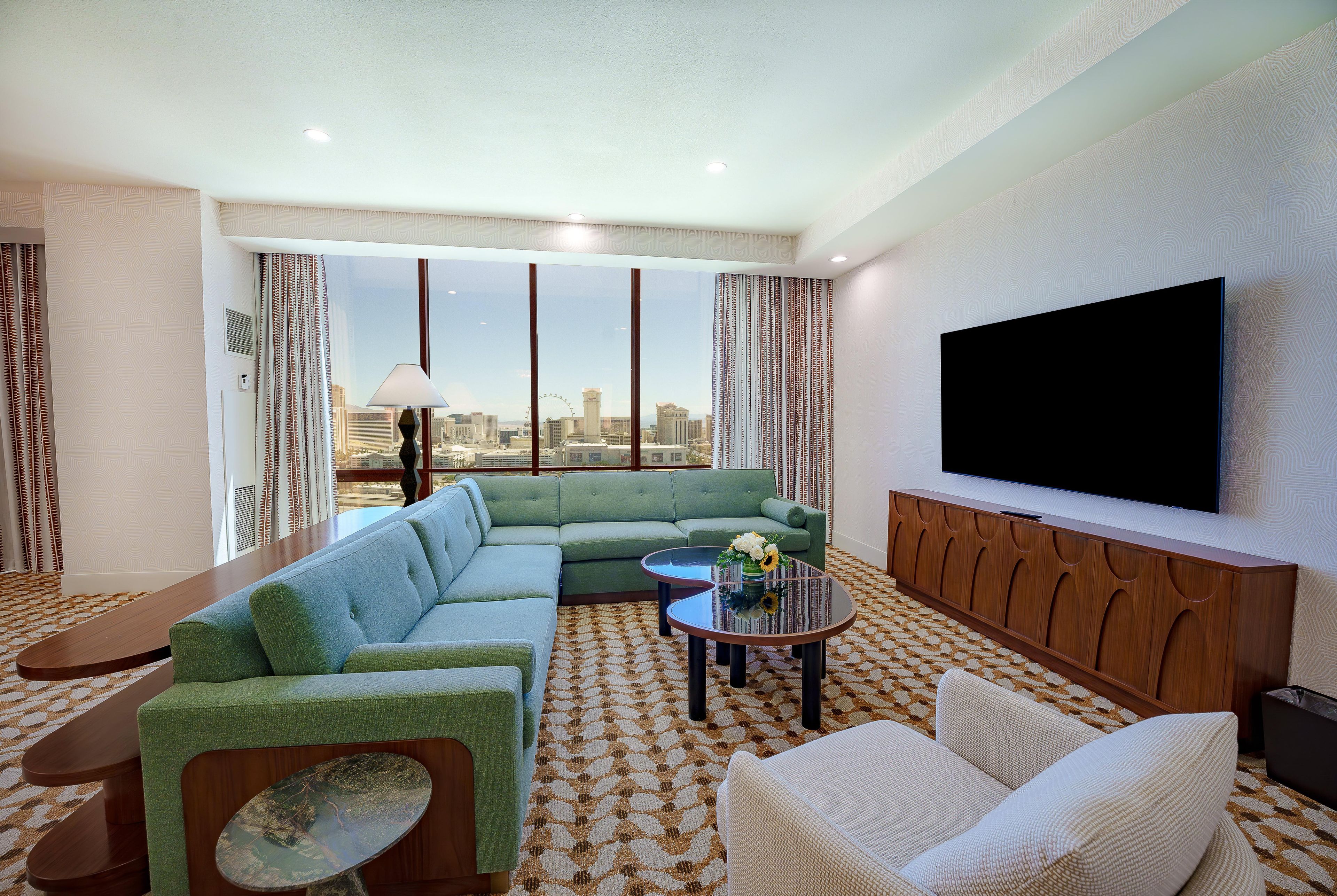 A modern living room with a sectional sofa, a small coffee table, a TV mounted on the wall, and a city view through large windows.