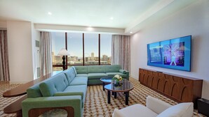 Ein modernes Hotelzimmer mit einem grünen Sofa, einem weißen Sessel, einem kleinen Couchtisch mit einer Vase und einem großen Fernseher an der Wand.