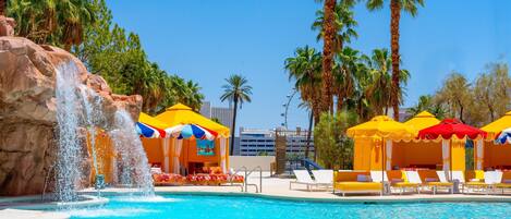 A pool with a waterfall feature, surrounded by palm trees and yellow umbrellas.