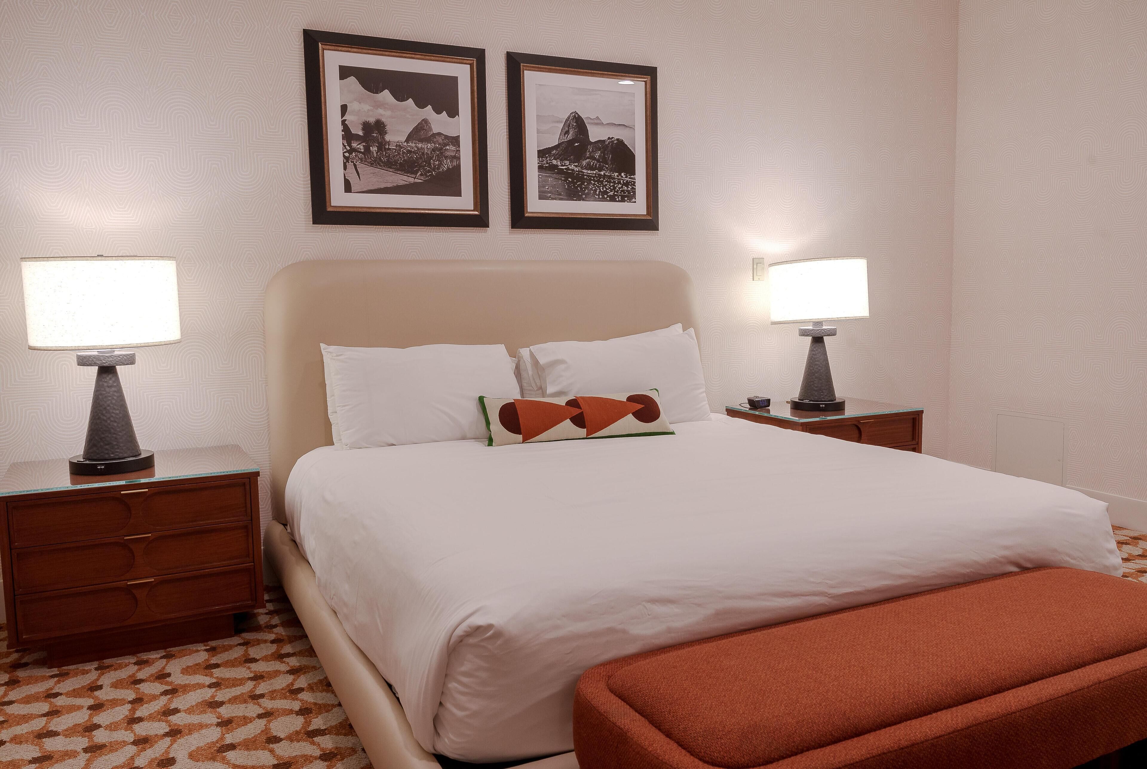 Habitación de hotel ordenada con una cama, dos mesitas de noche con lámparas, una mesita auxiliar y dos cuadros en la pared.