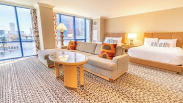 A hotel room with a large bed, a sofa, a small table, and a view of the cityscape through large windows.