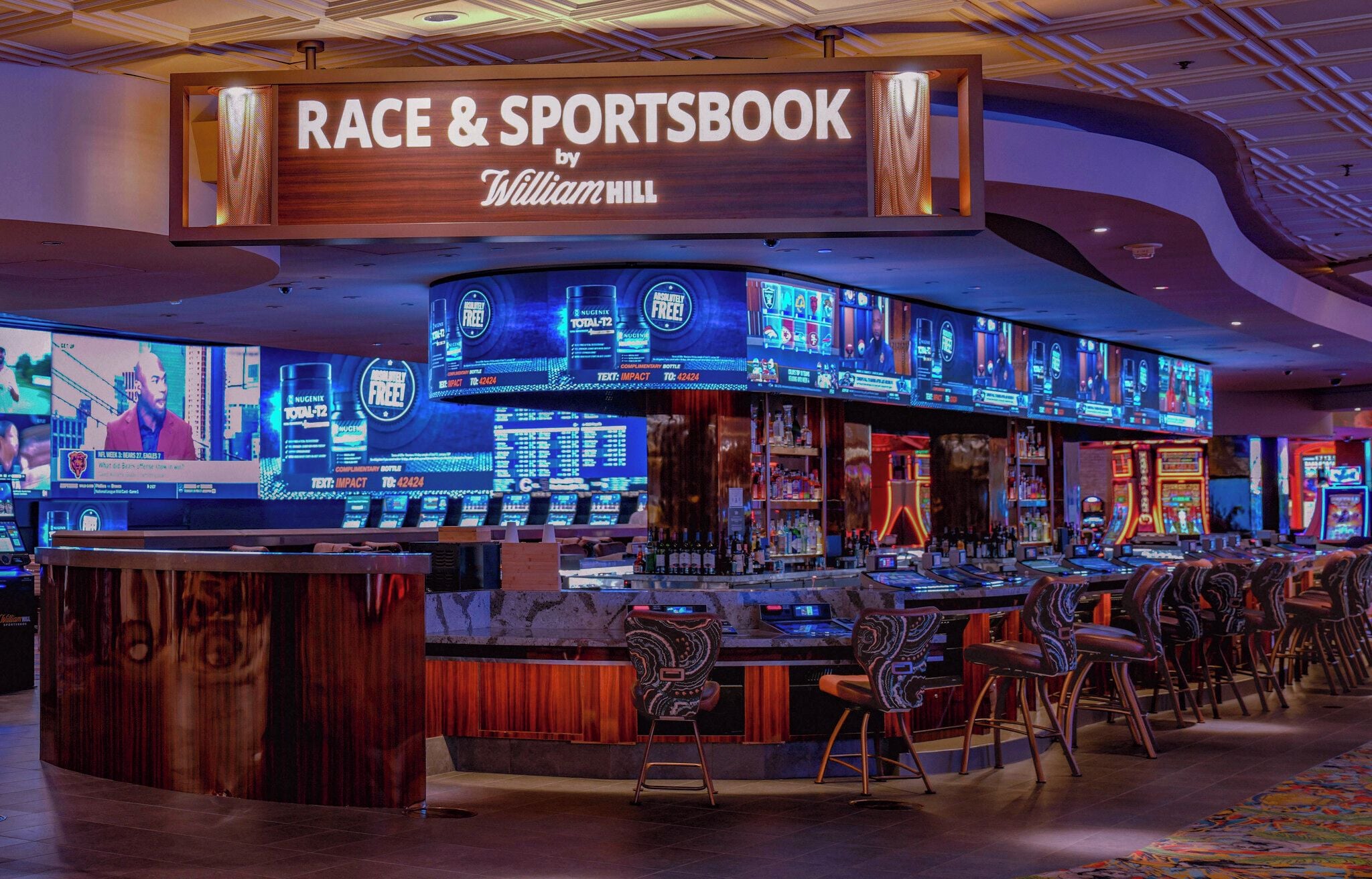 A bar area with a sign reading 'RACE & SPORTSBOOK by William Hill,' multiple screens displaying sports content, and a variety of seating options.