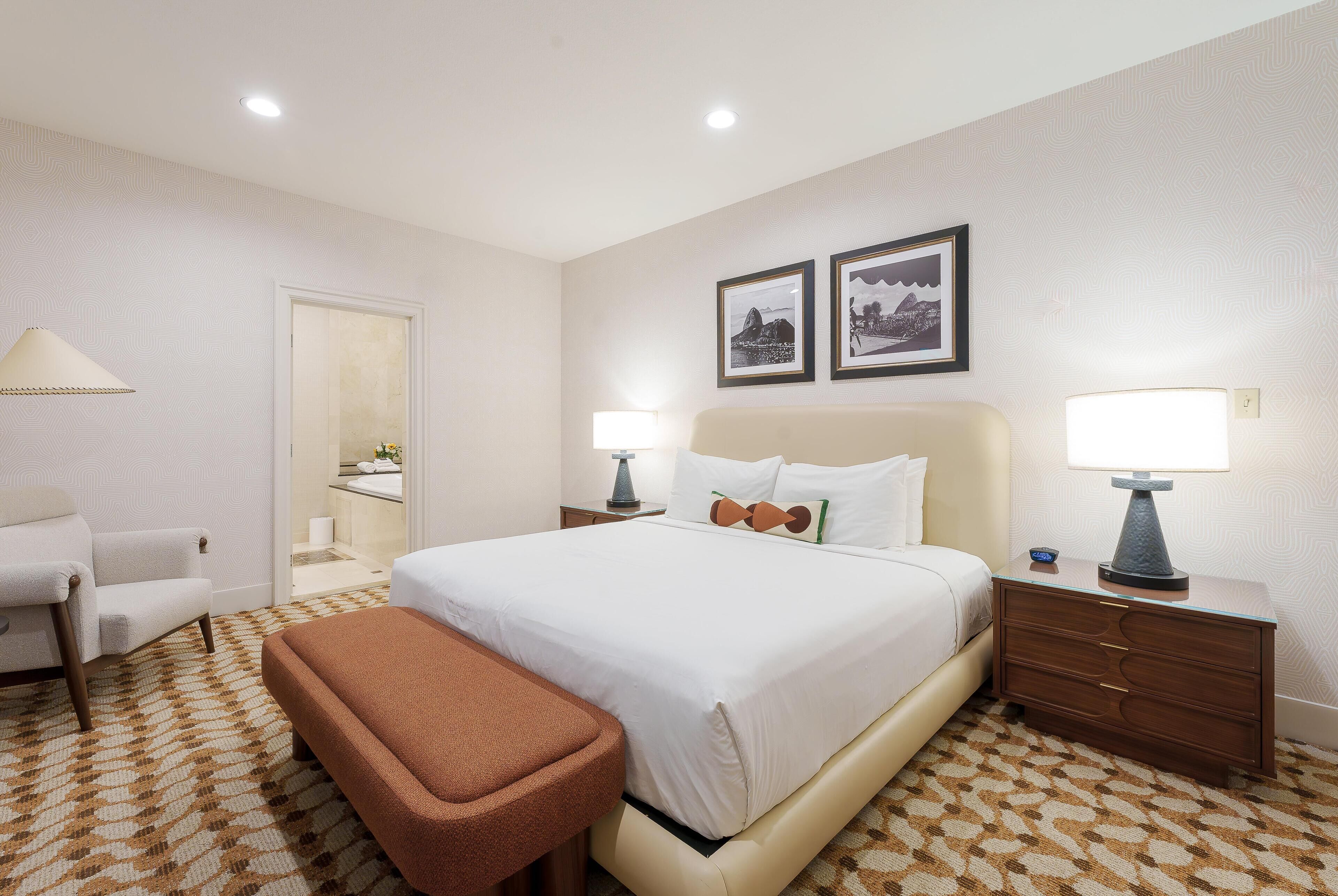 A hotel room with a large bed, two bedside lamps, a chair, a bench, and two framed pictures on the wall.