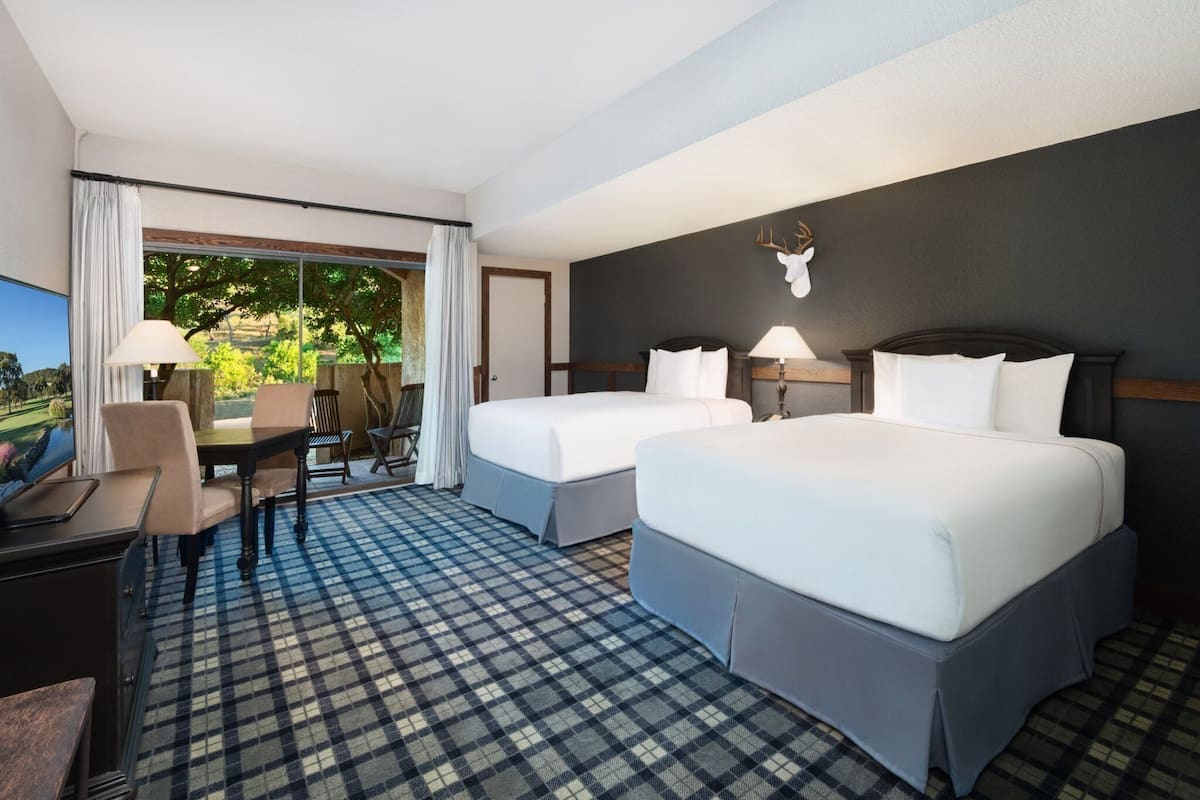 Une chambre d’hôtel avec deux lits, un bureau et une chaise. Une grande fenêtre offre une vue sur la verdure environnante.