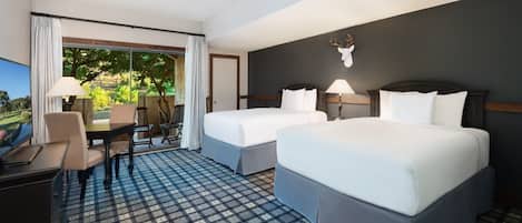A hotel room with two beds, a desk, and a chair. There is a large window with a view of greenery outside.