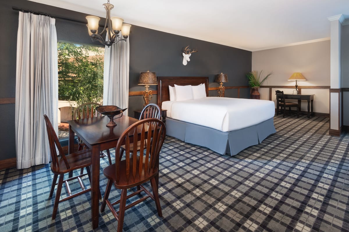 Une chambre d’hôtel avec un grand lit, une table à manger avec des chaises, une petite table avec une lampe, et une vue sur les arbres par la fenêtre.