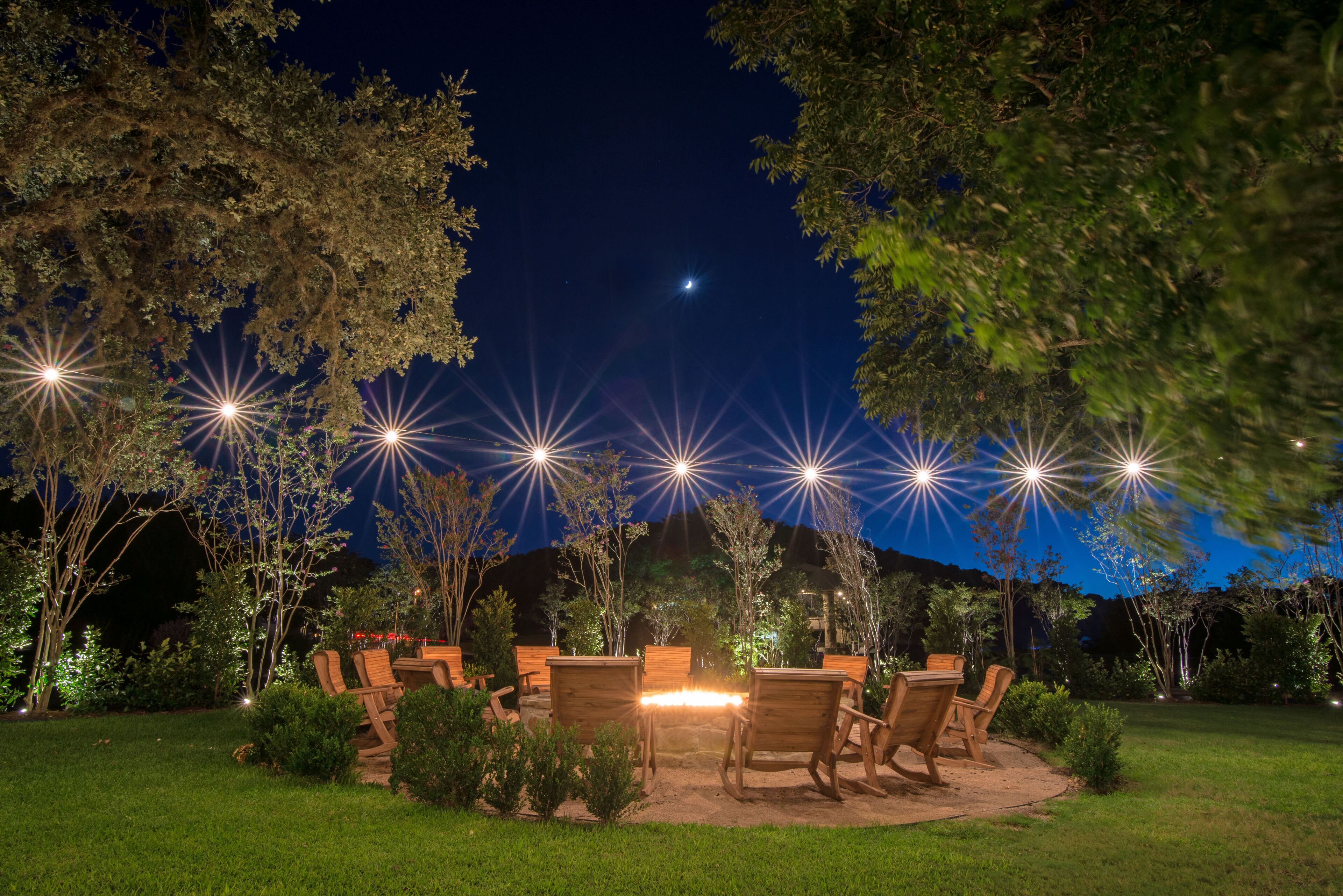 An outdoor seating area with wooden chairs and a table, surrounded by trees and illuminated by multiple light sources.
