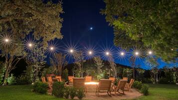 An outdoor seating area with wooden chairs and a table, surrounded by trees and illuminated by multiple light sources.