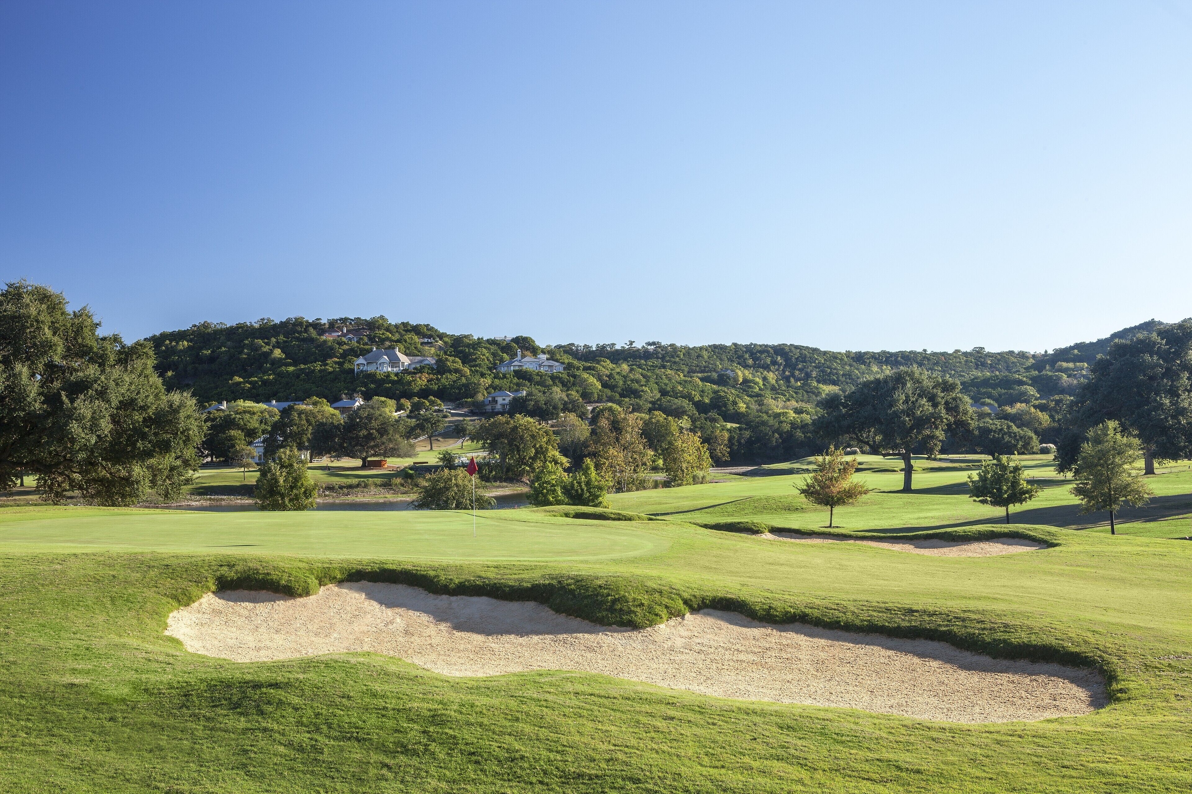 A golf course with sand bunkers, trees, and a hill with houses in the background.