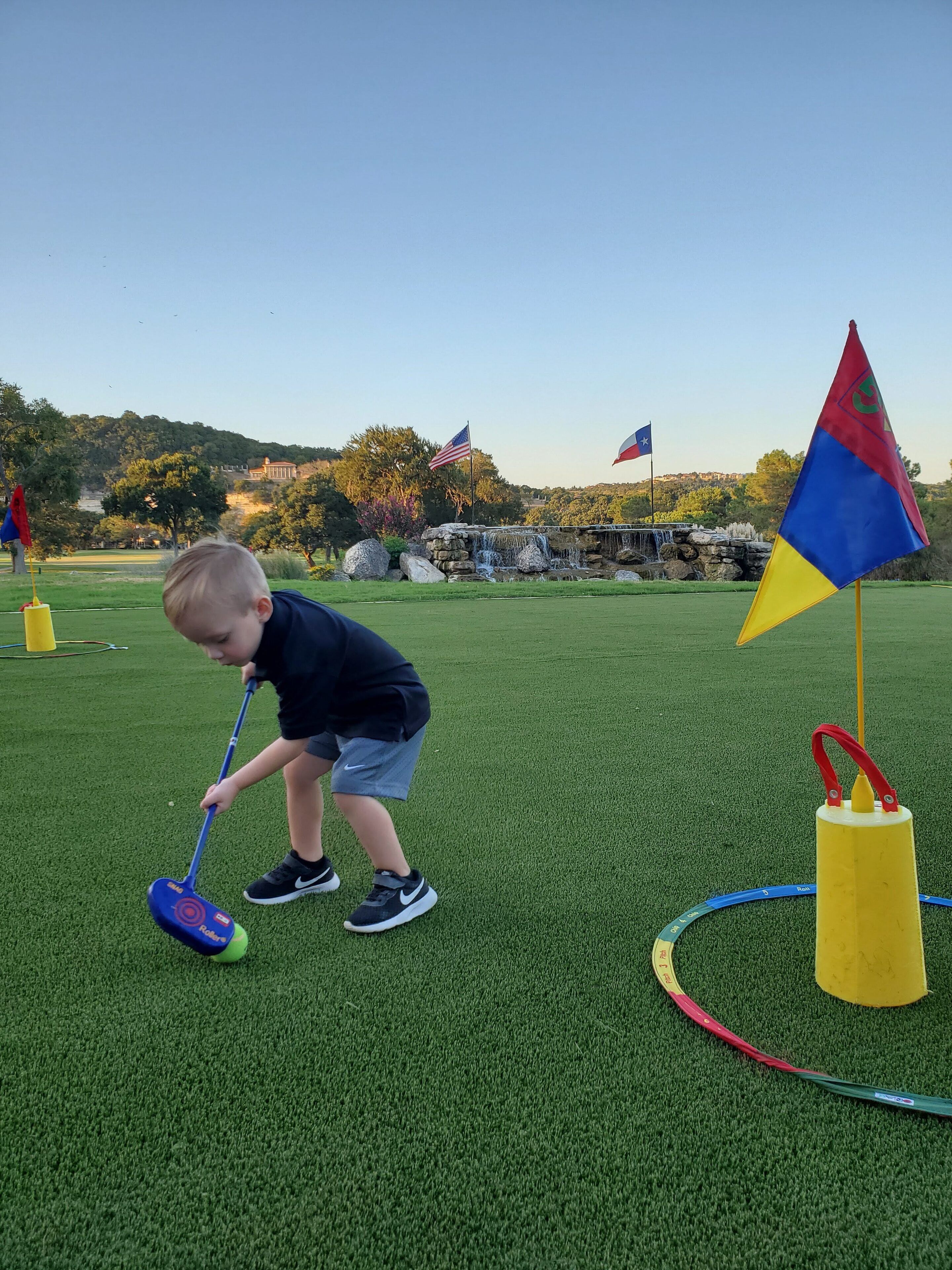 Un niño jugando con un juego de golf de juguete sobre césped artificial, con banderas y una fuente al fondo.