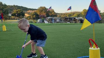 Un niño jugando con un juego de golf de juguete sobre césped artificial, con banderas y una fuente al fondo.