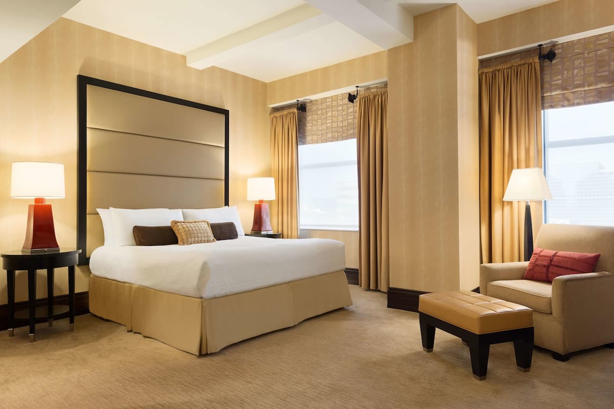 Quarto de hotel com uma cama grande, duas mesas de cabeceira com abajures, uma cadeira e uma janela com cortinas.