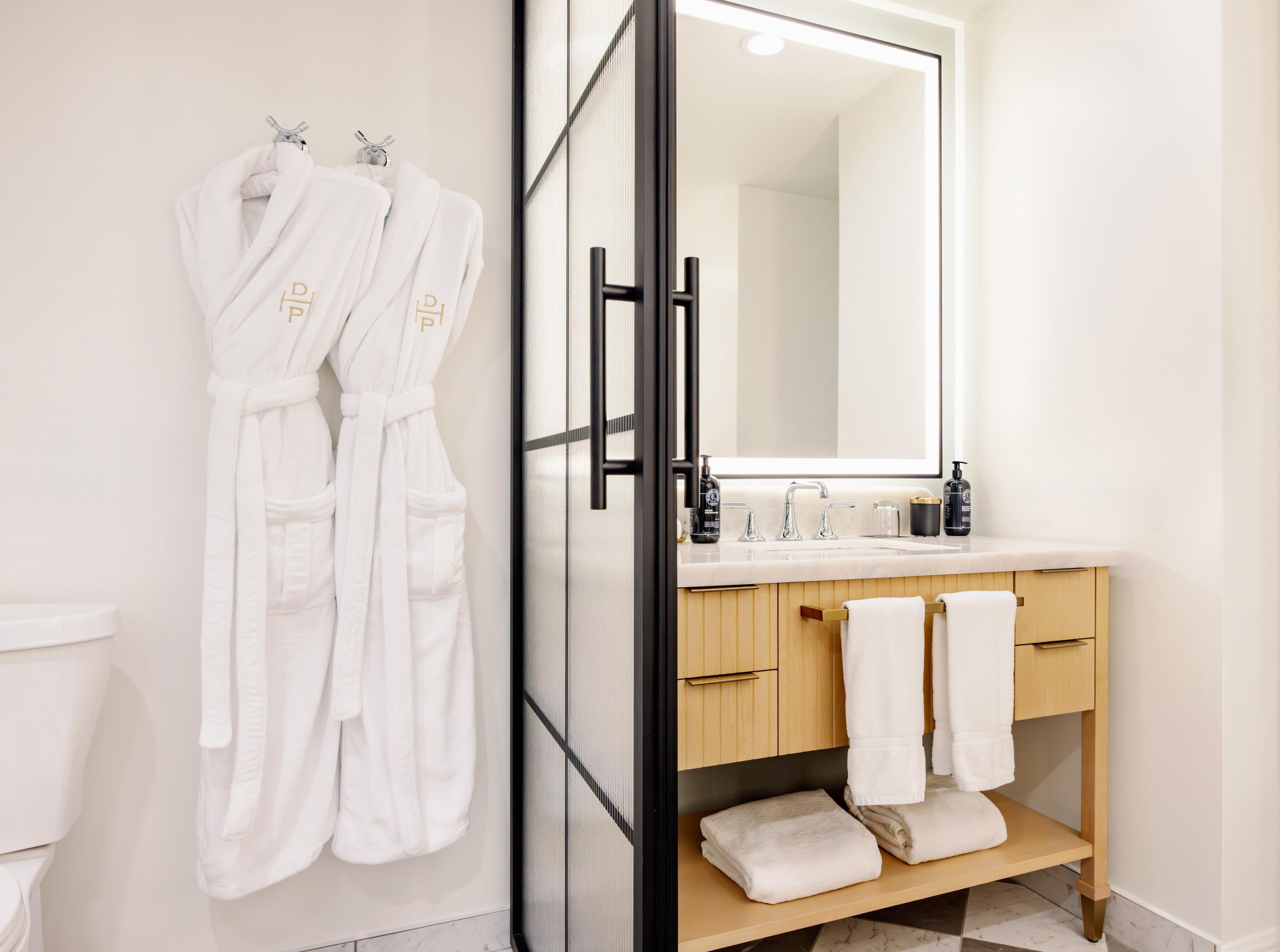 A modern bathroom with a glass shower door, a wooden vanity with a sink, and two white robes hanging on hooks.
