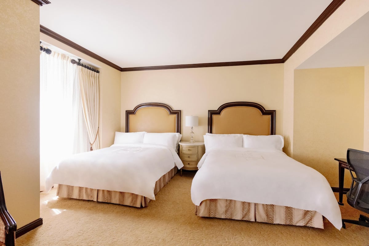 Quarto de hotel com duas camas, uma mesa de cabeceira com abajur, uma cadeira e uma janela com cortinas.