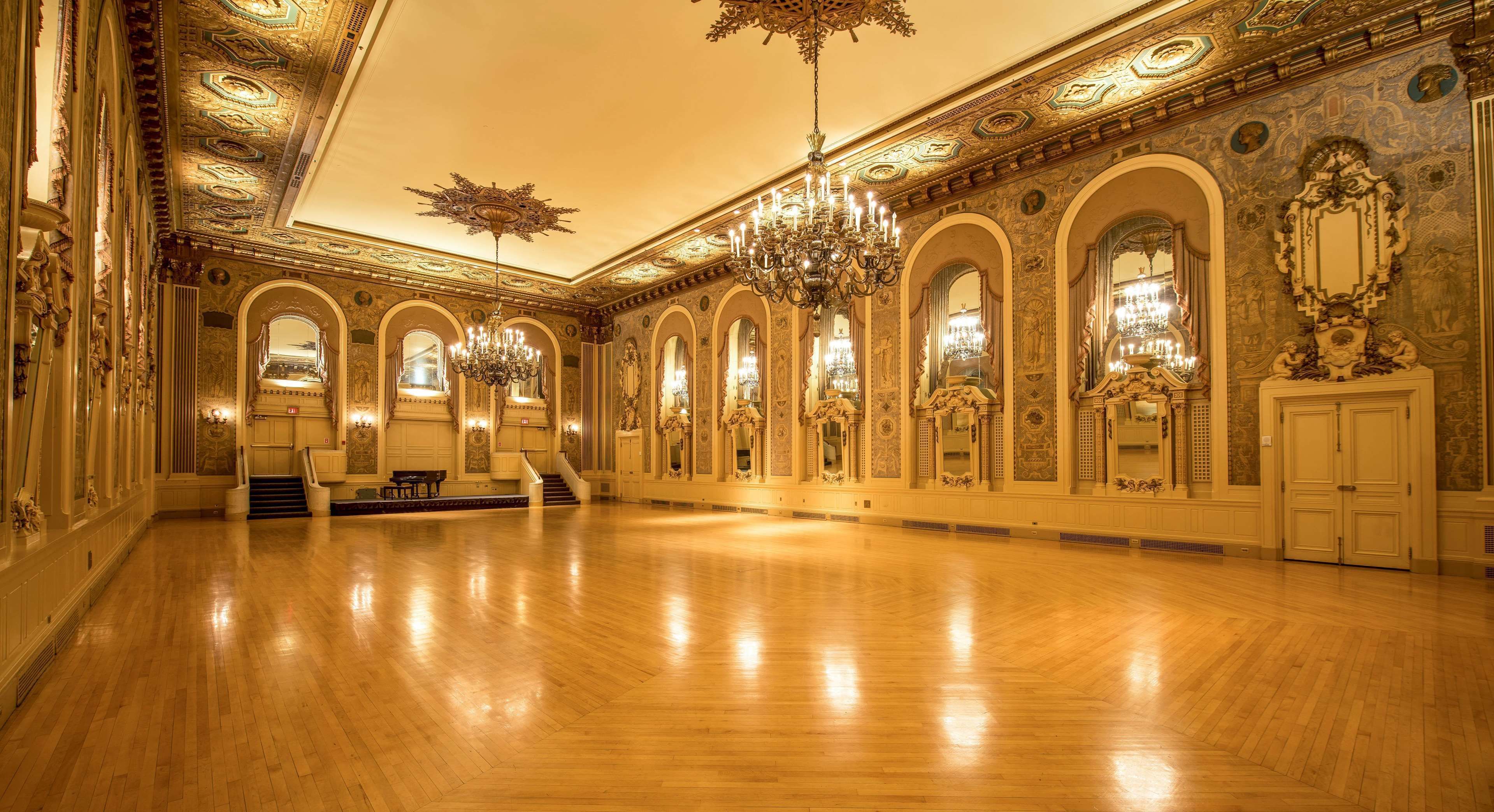 A spacious ballroom with ornate chandeliers, arched doorways, and detailed wall decorations.