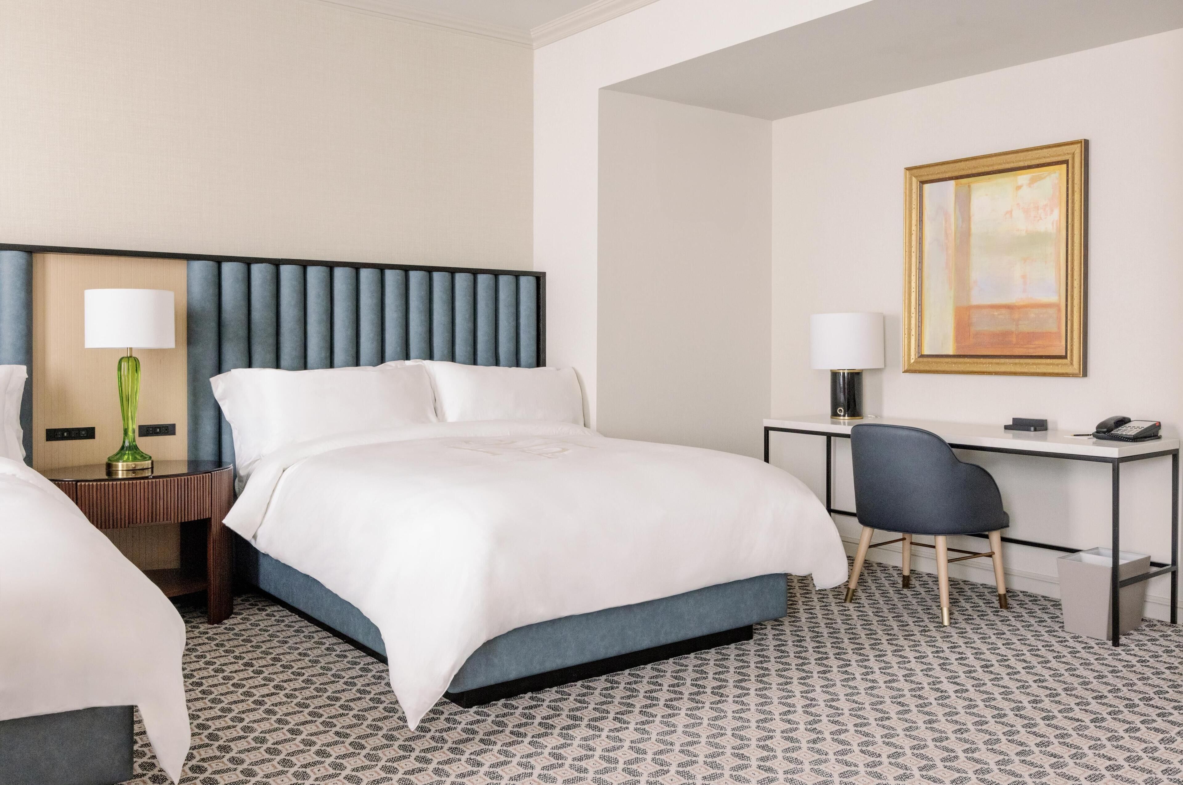 A hotel room with a bed, a desk, a chair, and a lamp.