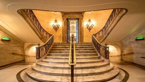 Una imponente escalera de mármol con barandillas de hierro ornamentadas y un pasamanos central, que conduce a una puerta con una araña colgante.