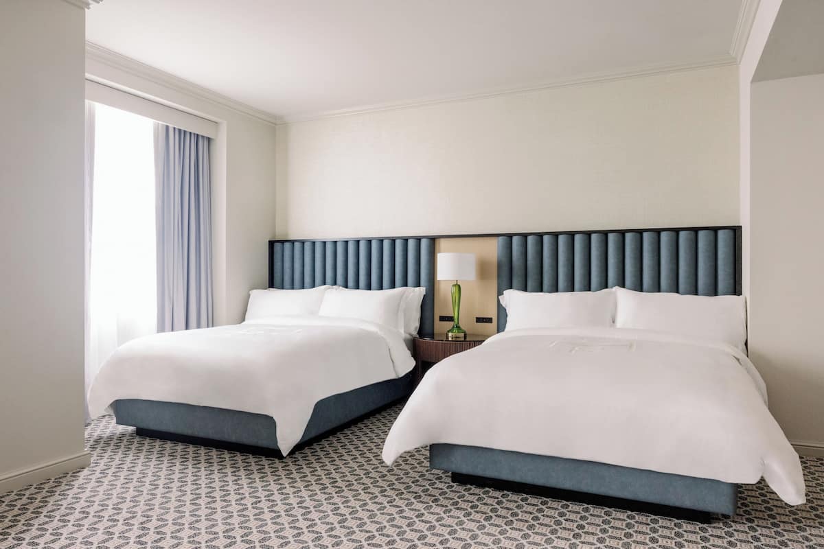 Quarto de hotel com duas camas, piso estampado, janela com cortinas e mesa de cabeceira com abajur.