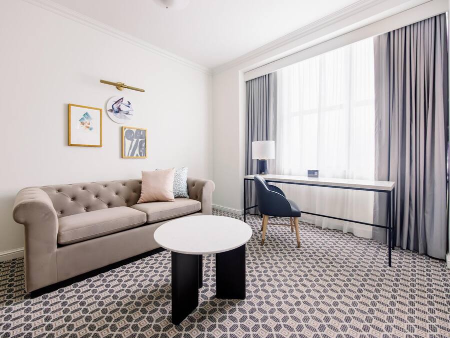 Een kamer met een bank, een ronde tafel, een bureau en een groot raam met gordijnen.