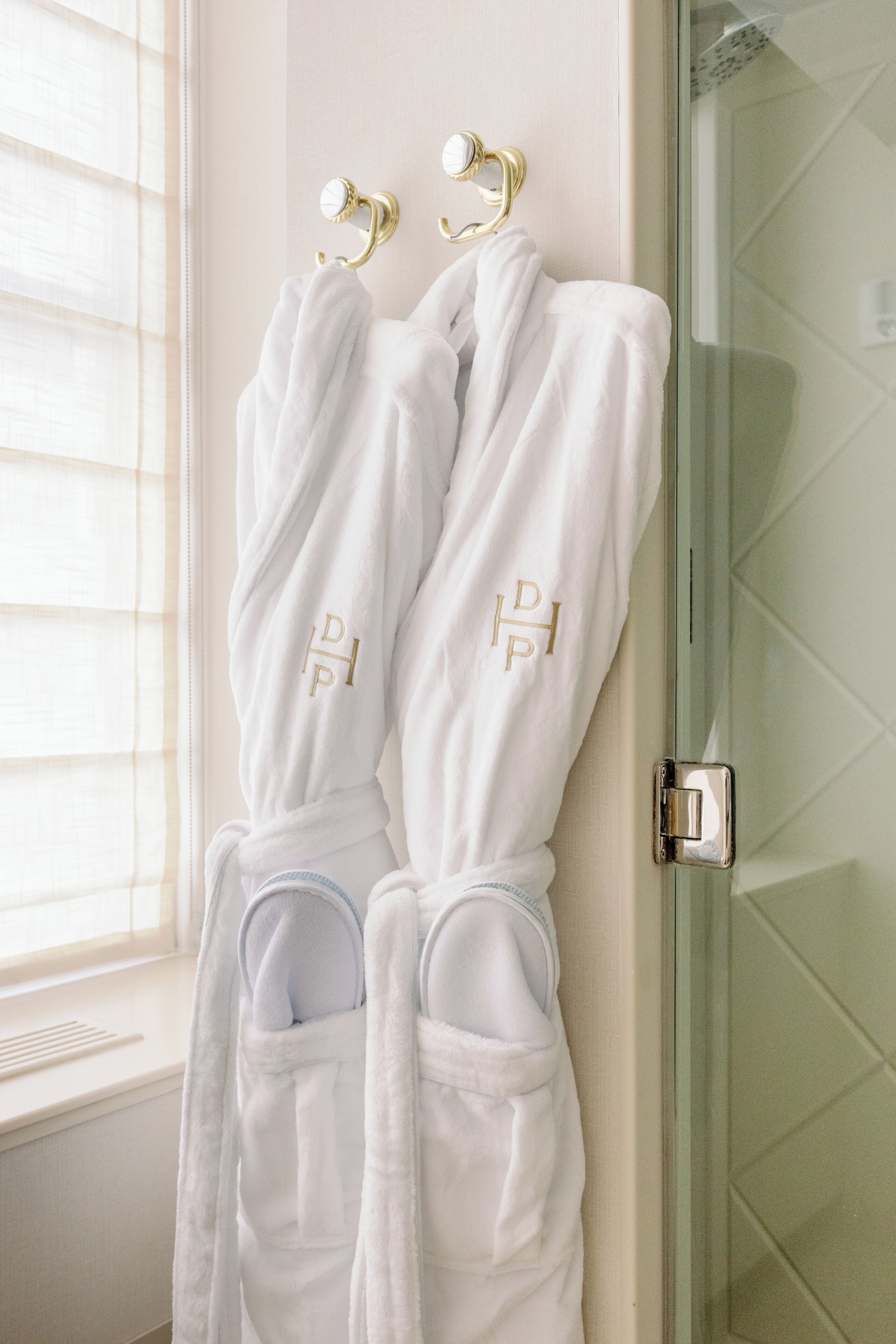 A bathroom with two white bathrobes hanging on hooks, a glass shower door, and a window with blinds.