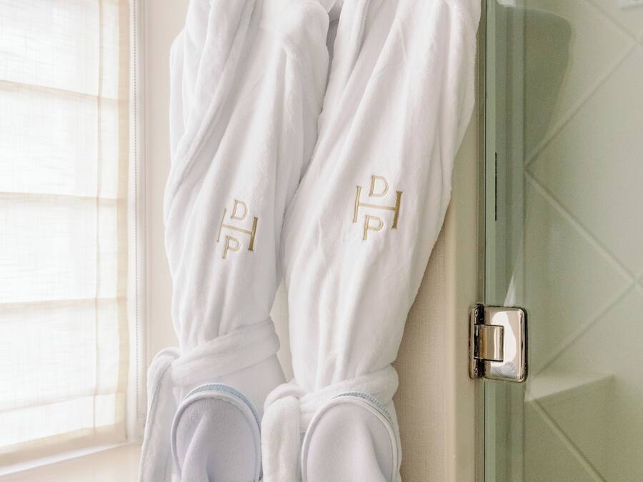 A bathroom with two white bathrobes hanging on hooks, a glass shower door, and a window with blinds.