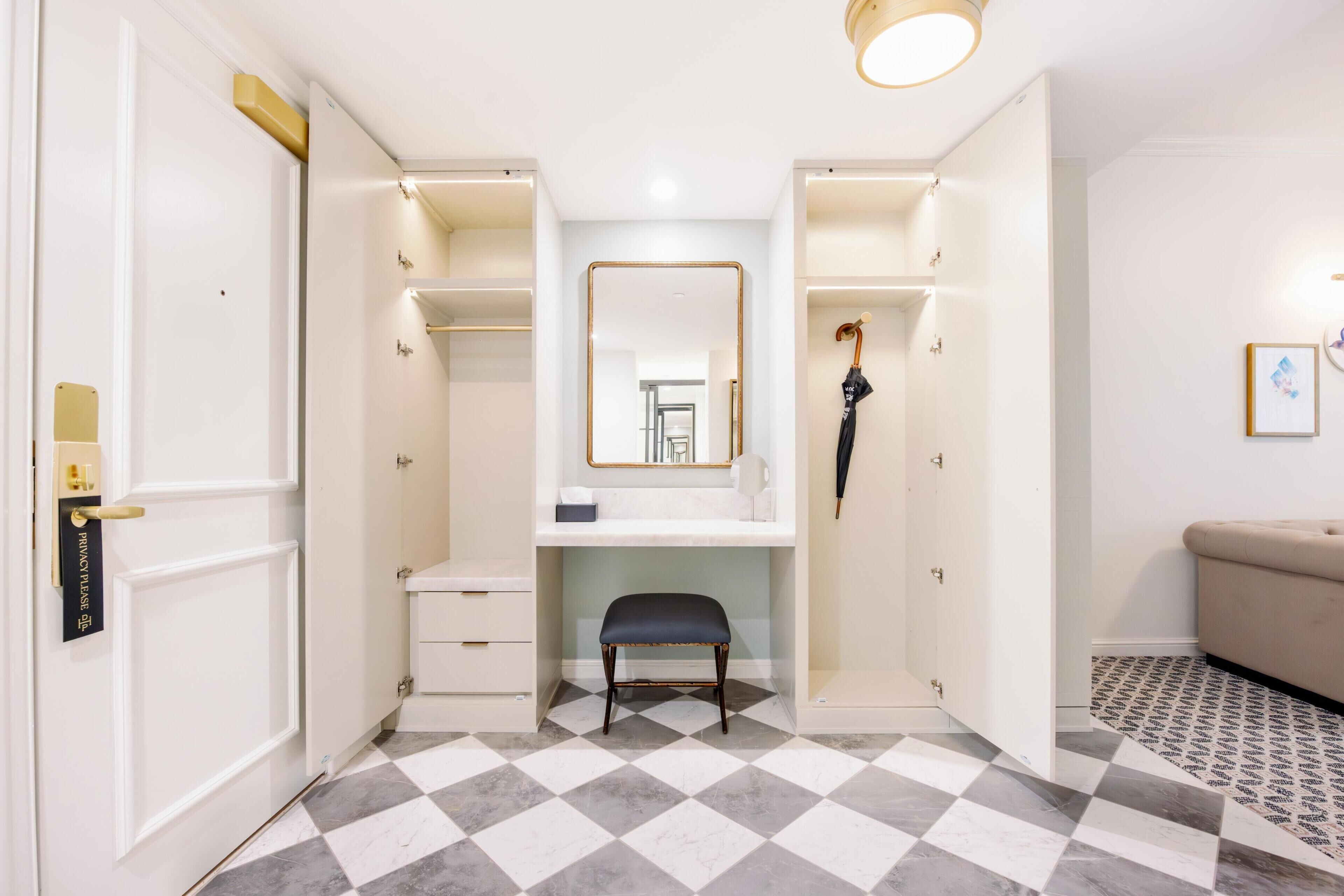 A hotel room with a checkered floor, a desk with a mirror, a bench, and a wardrobe.