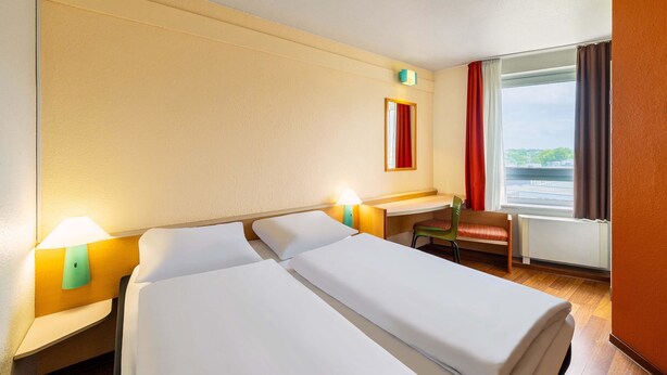 A hotel room with two beds, a desk, a chair, a window with curtains, and a lamp. - B&B HOTEL Bochum Hbf-Süd (Bochum)