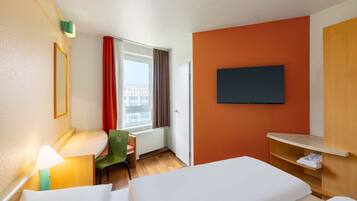 A hotel room with a bed, a desk with a chair, a TV mounted on the wall, and a window with curtains.